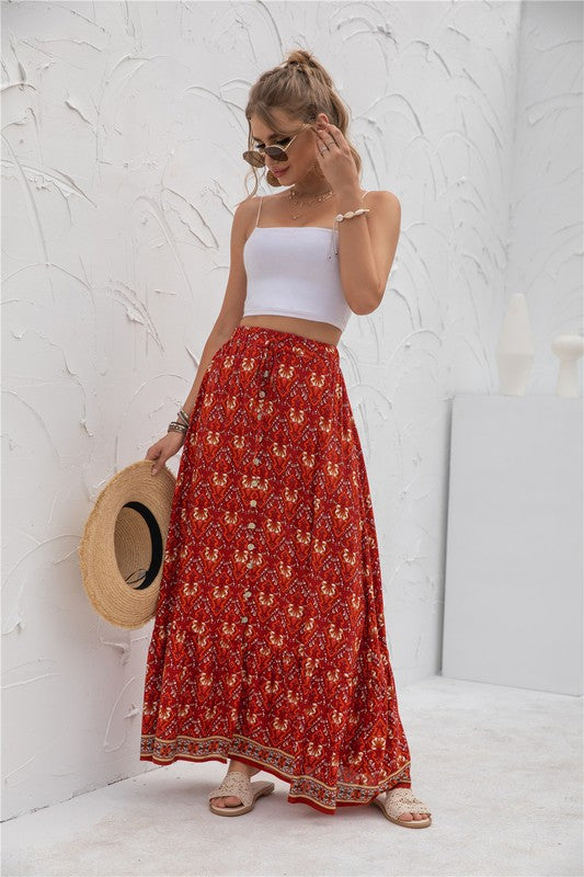 A woman stands against a light-textured wall wearing a white crop top, sunglasses, and holding a woven hat. She complements her look with the Womens Print Maxi Skirt, an eye-catching long red skirt with patterns.