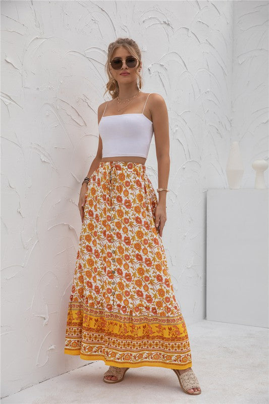 A woman stands against a light-textured wall wearing a white crop top, sunglasses, and holding a woven hat. She complements her look with the Womens Print Maxi Skirt, an eye-catching long red skirt with patterns.