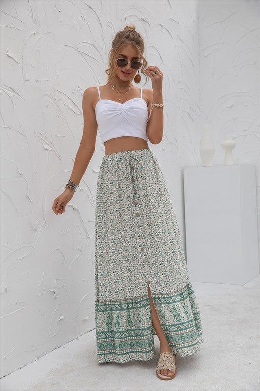 A woman stands against a light-textured wall wearing a white crop top, sunglasses, and holding a woven hat. She complements her look with the Womens Print Maxi Skirt, an eye-catching long red skirt with patterns.