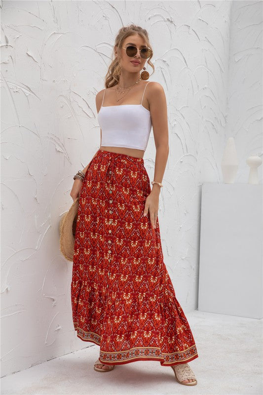 A woman stands against a light-textured wall wearing a white crop top, sunglasses, and holding a woven hat. She complements her look with the Womens Print Maxi Skirt, an eye-catching long red skirt with patterns.