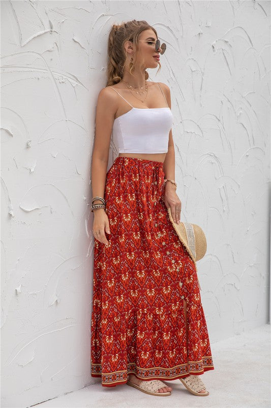 A woman stands against a light-textured wall wearing a white crop top, sunglasses, and holding a woven hat. She complements her look with the Womens Print Maxi Skirt, an eye-catching long red skirt with patterns.