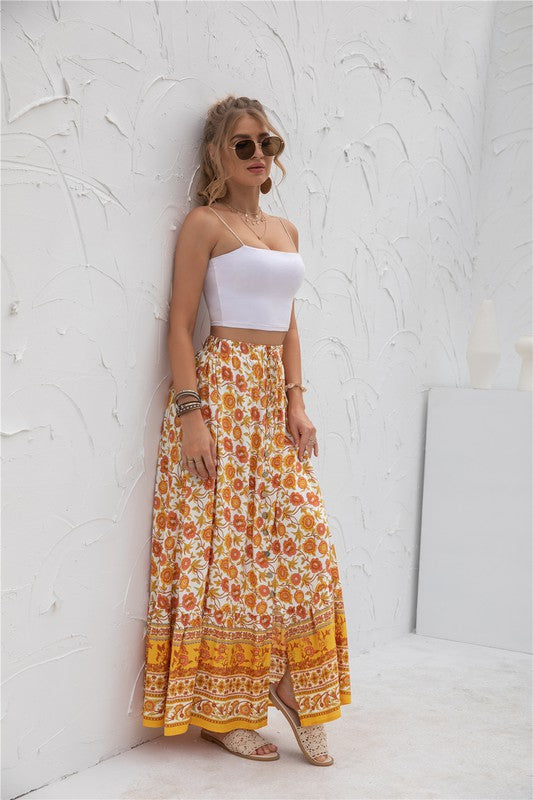 A woman stands against a light-textured wall wearing a white crop top, sunglasses, and holding a woven hat. She complements her look with the Womens Print Maxi Skirt, an eye-catching long red skirt with patterns.