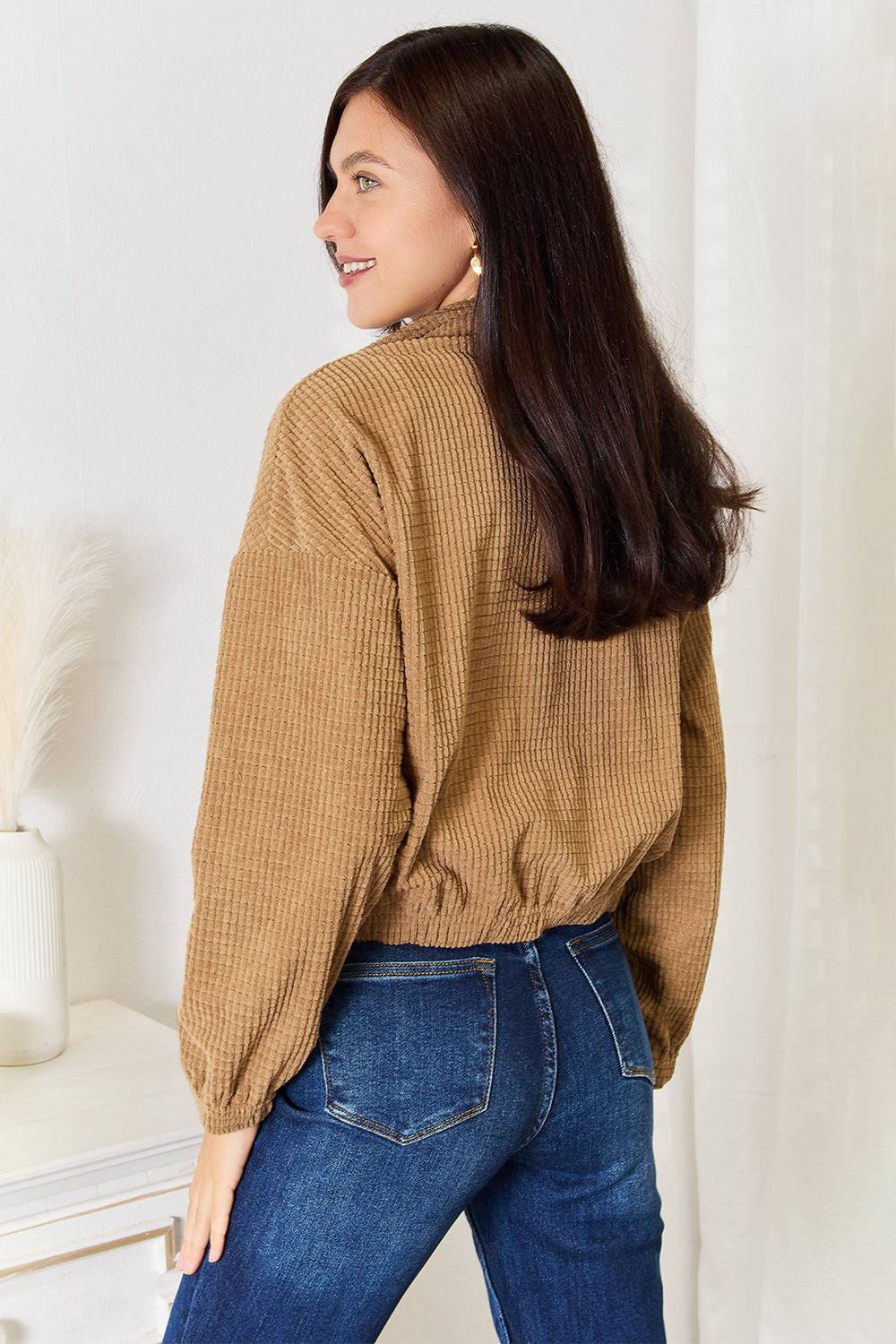 A person wearing the versatile Perfee Long Sleeve Dropped Shoulder Jacket in brown corduroy and blue jeans is standing indoors, smiling, with one hand resting on a white piece of furniture.