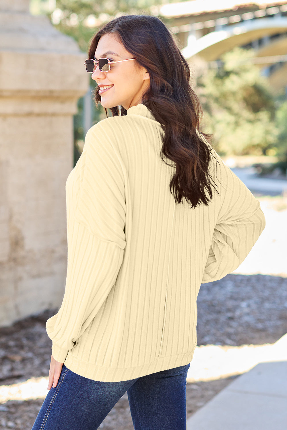 A woman with long hair is wearing a green Basic Bae Full Size Ribbed Exposed Seam Mock Neck Knit Top, blue jeans, and sunglasses. She is standing outdoors and smiling.