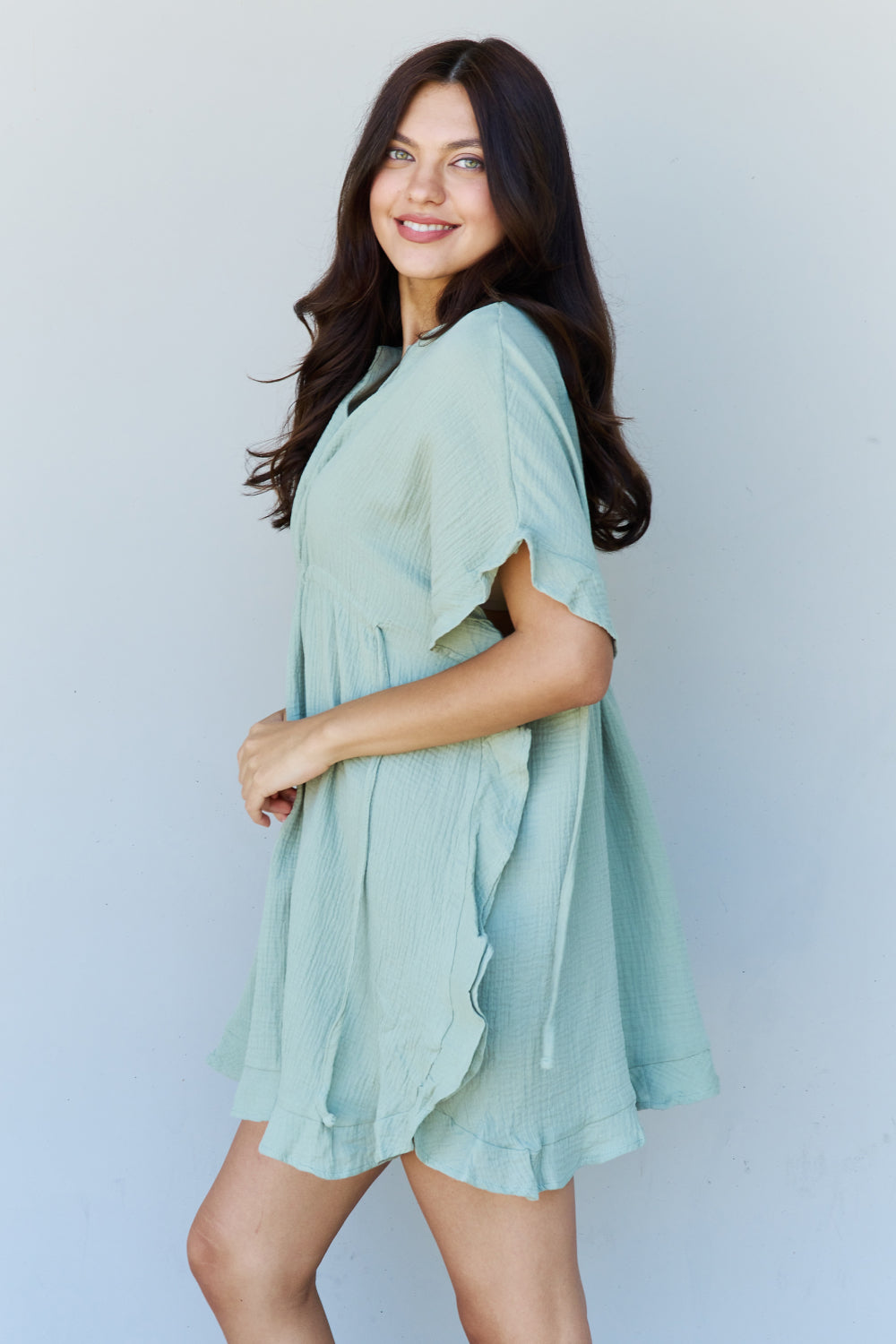 A woman is wearing the Ninexis Out Of Time Full Size Ruffle Hem Dress with Drawstring Waistband in Light Sage. Her long, dark hair falls gracefully as she smiles and looks to her left, standing against a plain background.