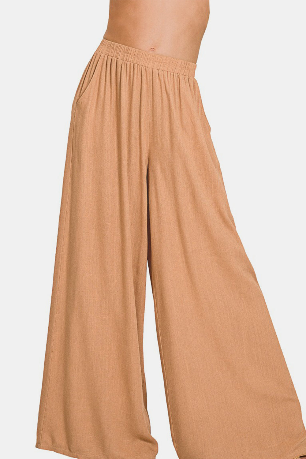 A person wearing Zenana Pleated Linen Blend Wide Leg Pants in tan, with hands in pockets, standing against a plain white background. Upper body and face are not visible.