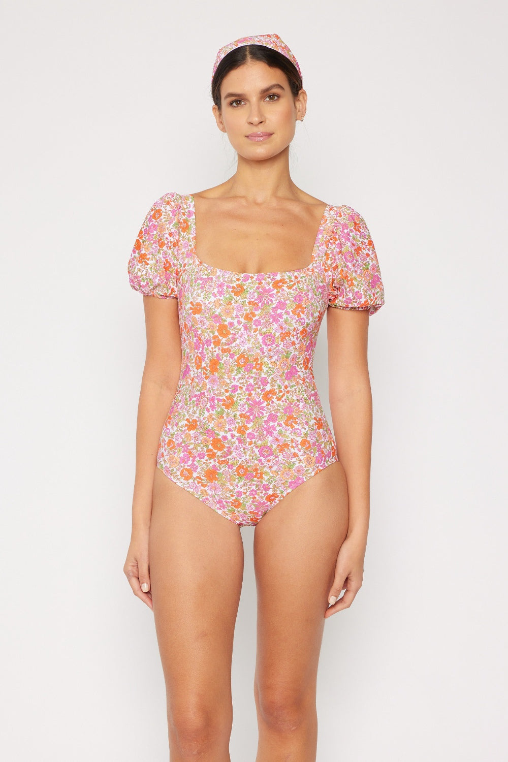 A woman poses in the Marina West Swim Floral Puff Sleeve One-Piece swimsuit, which features removable pads and comes with a matching headband, set against a plain background.