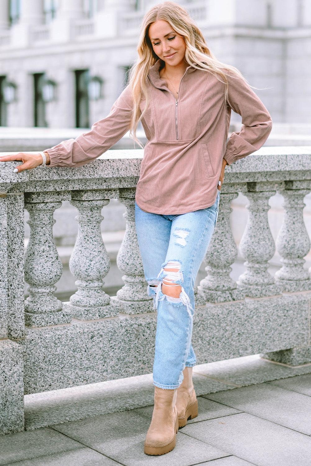 A person with long blonde hair, wearing a trendy Apricot Ribbed Texture Half Zip Collared Sweatshirt and blue jeans, stands facing away and looks over a stone balustrade.