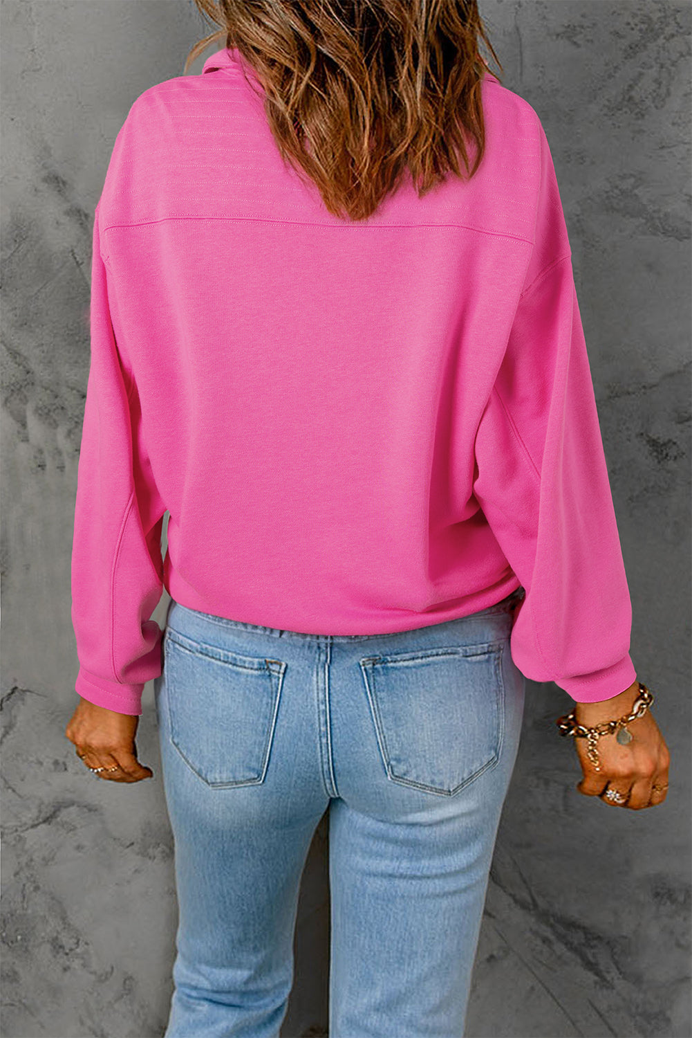 A person with long hair is seen from behind, wearing a Bright Pink Vintage Mineral Wash Half Button Collared Sweatshirt and light blue jeans.