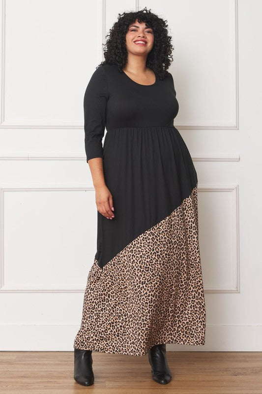 A woman with curly hair is wearing the Asymmetrical Leopard Accent Maxi Dress, featuring a long black design with a floral patterned section at the bottom. She stands against a white paneled wall, her hands tucked in her dress pockets, showcasing its elegant design.