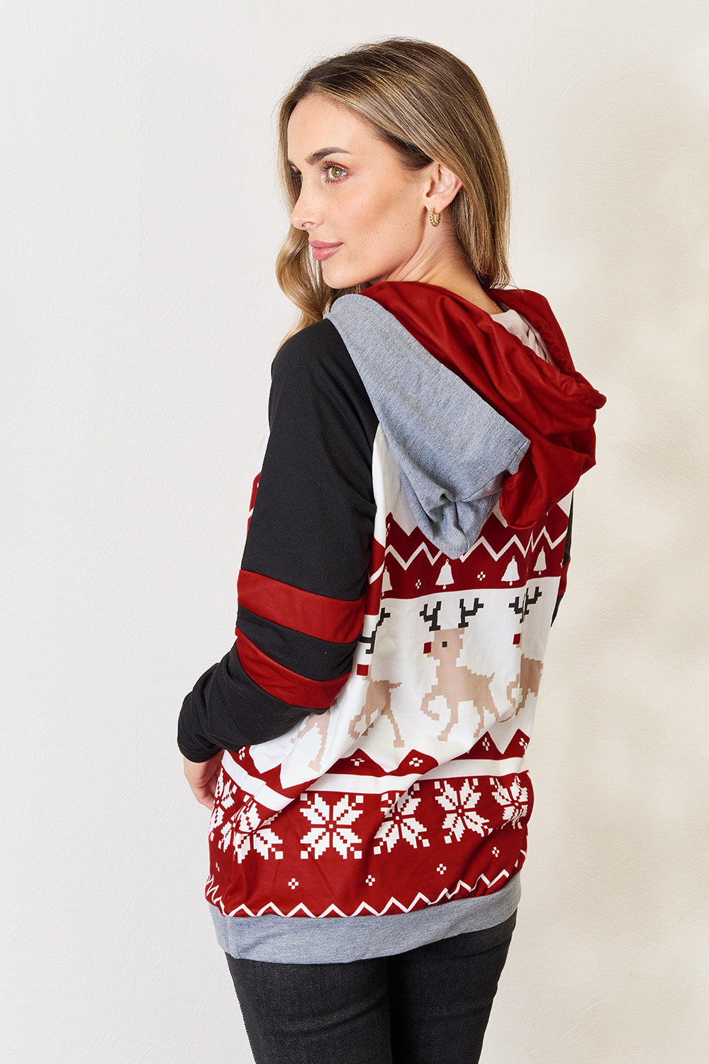 A person is wearing the Double Take Full Size Christmas Drawstring Long Sleeve Hoodie, which features a holiday-themed red and white pattern with reindeer and snowflakes. This cozy hoodie, made from a polyester spandex blend, has stylish black sleeves accented with red stripes. The person is smiling and looking to the side.