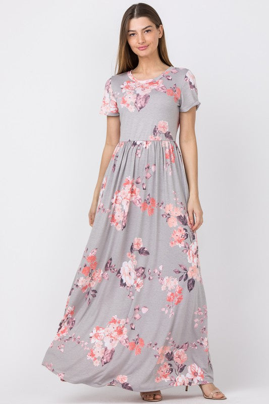 A woman wearing the Vintage Floral Maxi Dress With Pockets, showcasing a pink floral pattern on gray fabric, stands against a plain background. This elegant dress combines style with practicality, offering convenient pockets for added functionality.