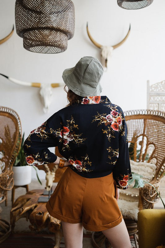 A woman wearing the Lush Rose Kimono, brown shorts, and a wide-brimmed hat stands indoors, in front of various decorative items and plants, looking directly at the camera.