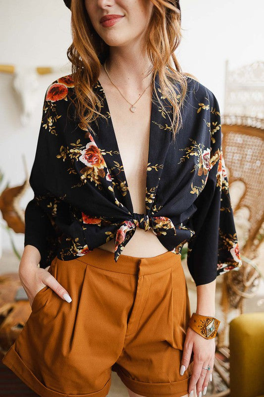 A woman wearing the Lush Rose Kimono, brown shorts, and a wide-brimmed hat stands indoors, in front of various decorative items and plants, looking directly at the camera.