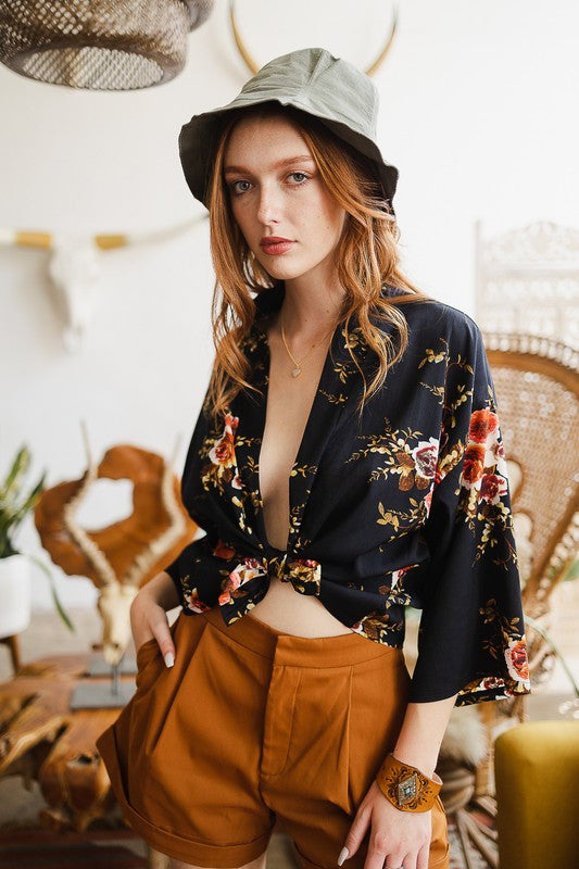 A woman wearing the Lush Rose Kimono, brown shorts, and a wide-brimmed hat stands indoors, in front of various decorative items and plants, looking directly at the camera.