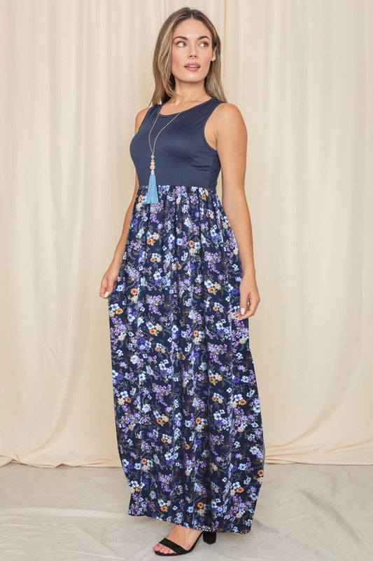 A woman wearing a Sleeveless Floral Maxi Dress leans against a light beige curtain. Her navy polyester dress pairs perfectly with her black sandals, and she complements the look with a necklace adorned with a blue tassel.