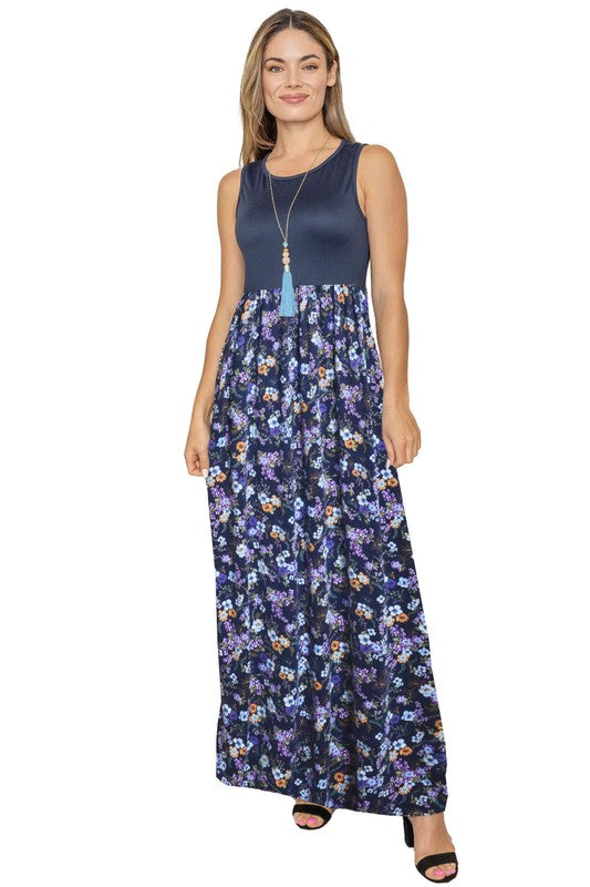 A woman wearing a Sleeveless Floral Maxi Dress leans against a light beige curtain. Her navy polyester dress pairs perfectly with her black sandals, and she complements the look with a necklace adorned with a blue tassel.