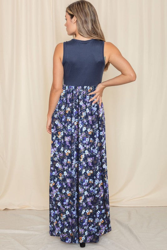 A woman wearing a Sleeveless Floral Maxi Dress leans against a light beige curtain. Her navy polyester dress pairs perfectly with her black sandals, and she complements the look with a necklace adorned with a blue tassel.