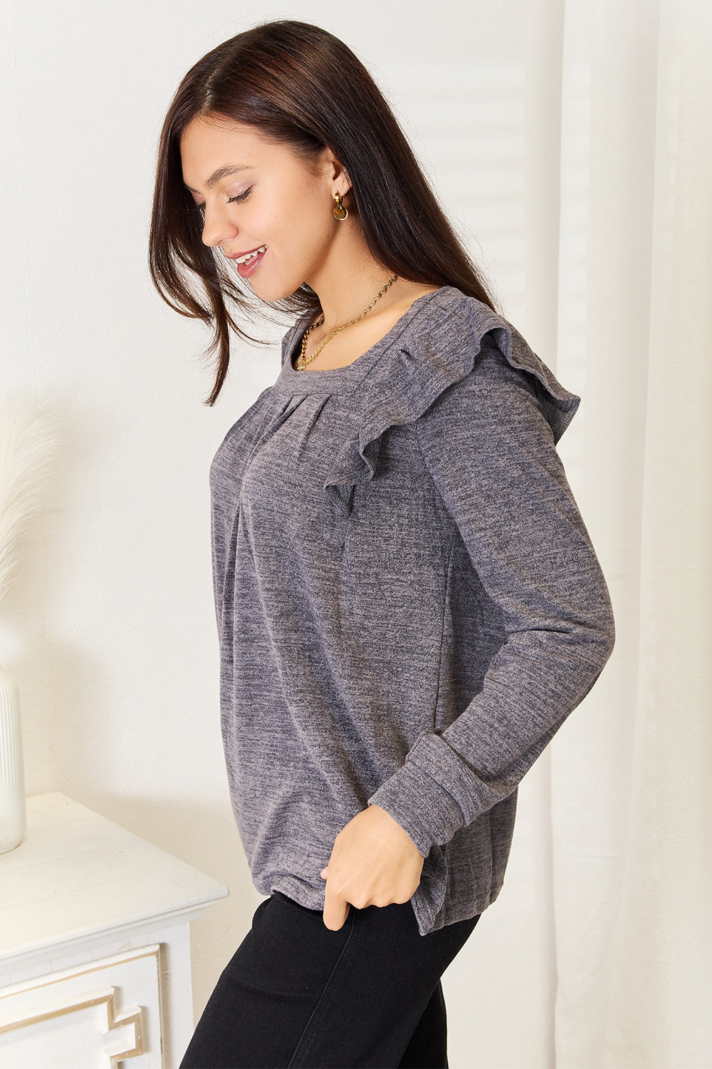 A woman wearing a Double Take Square Neck Ruffle Shoulder Long Sleeve T-Shirt in pink and blue jeans stands with one hand in her pocket, smiling. She has long curly hair and wears a necklace and bracelet. She stands in a room with white decor.