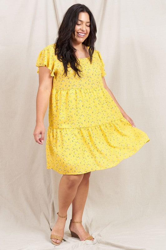 A person is standing and smiling, wearing the Plus Floral Flutter Sleeve Tunic, made from 100% polyester and holding a small wooden handbag. They are positioned against a neutral backdrop.