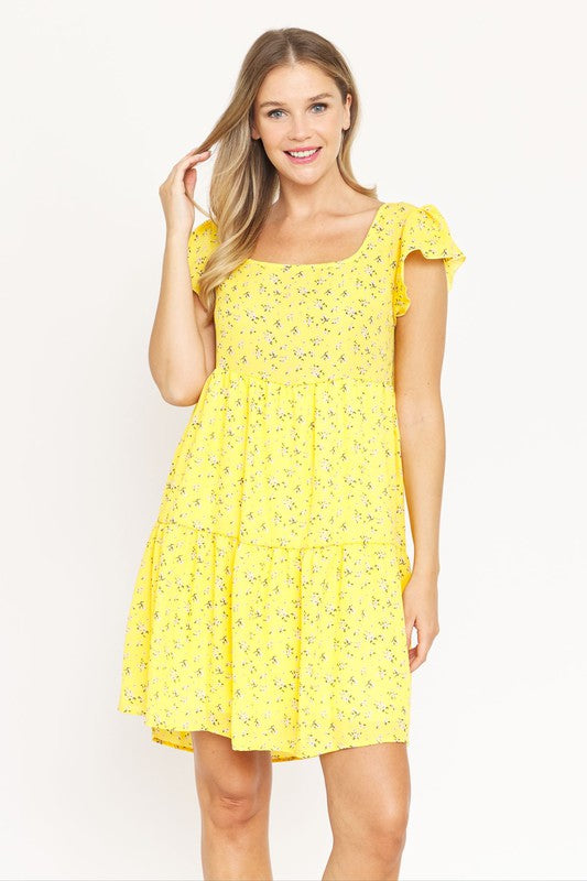 A person is standing and smiling, wearing the Plus Floral Flutter Sleeve Tunic, made from 100% polyester and holding a small wooden handbag. They are positioned against a neutral backdrop.