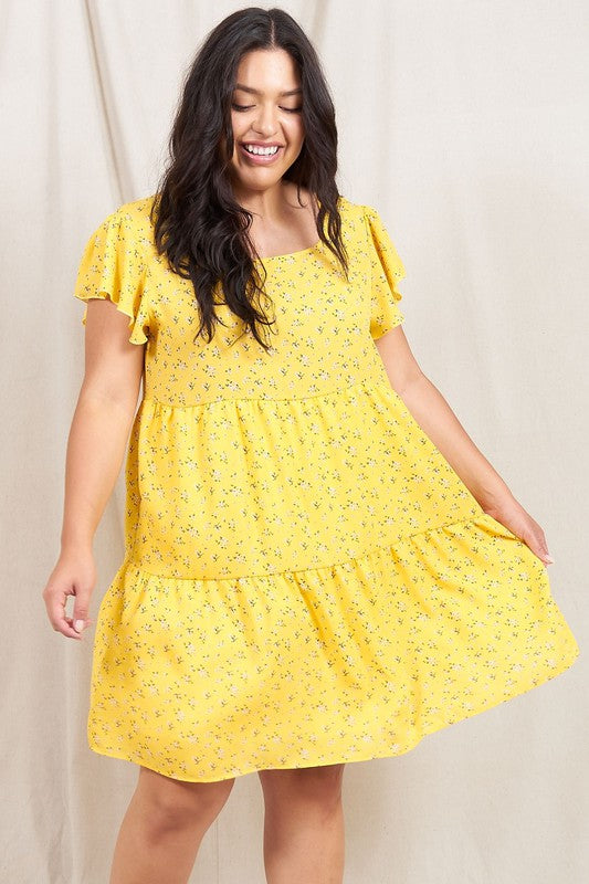 A person is standing and smiling, wearing the Plus Floral Flutter Sleeve Tunic, made from 100% polyester and holding a small wooden handbag. They are positioned against a neutral backdrop.