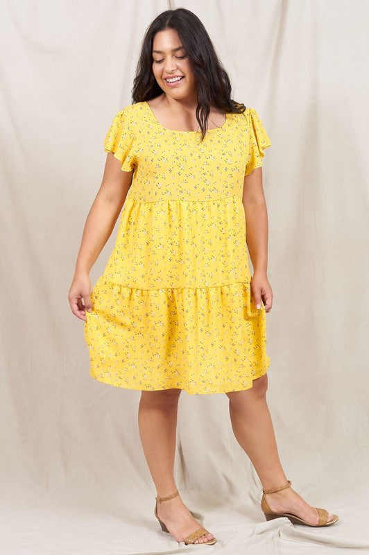 A person is standing and smiling, wearing the Plus Floral Flutter Sleeve Tunic, made from 100% polyester and holding a small wooden handbag. They are positioned against a neutral backdrop.