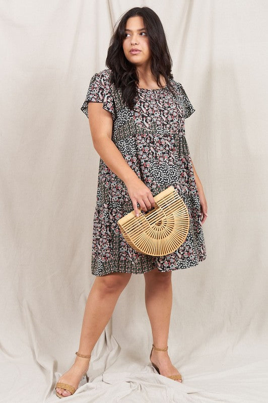 A person is standing and smiling, wearing the Plus Floral Flutter Sleeve Tunic, made from 100% polyester and holding a small wooden handbag. They are positioned against a neutral backdrop.