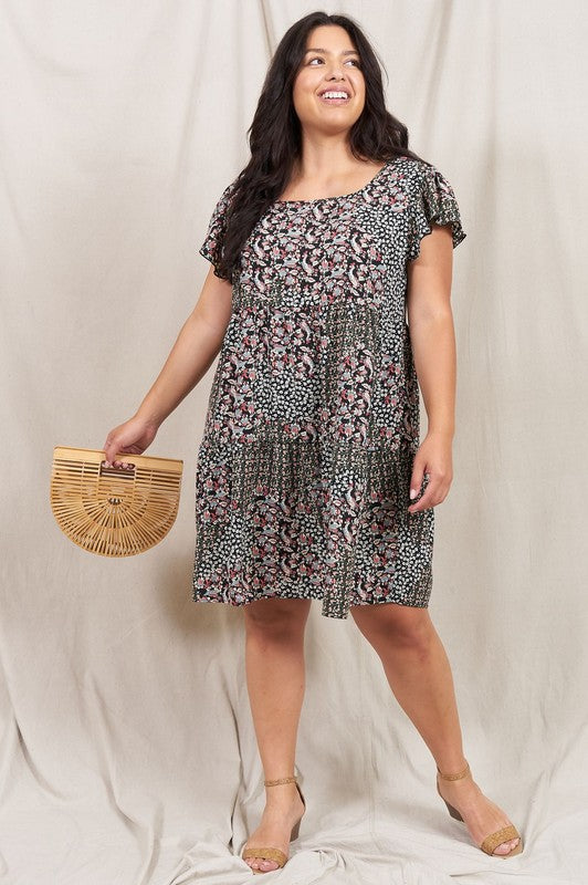 A person is standing and smiling, wearing the Plus Floral Flutter Sleeve Tunic, made from 100% polyester and holding a small wooden handbag. They are positioned against a neutral backdrop.