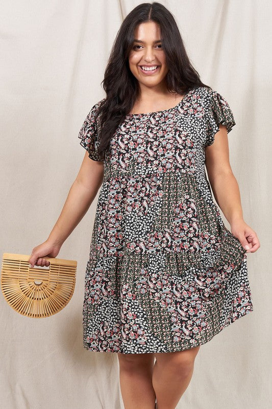 A person is standing and smiling, wearing the Plus Floral Flutter Sleeve Tunic, made from 100% polyester and holding a small wooden handbag. They are positioned against a neutral backdrop.