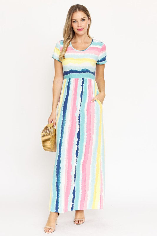 A woman stands wearing a vibrant Water Color Short Sleeve Maxi Dress, proudly made in the United States, and holding a woven handbag. One of her hands rests casually in the pocket of her dress.