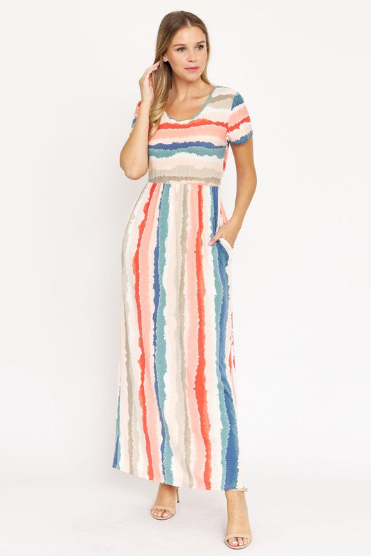 A woman stands against a beige backdrop, wearing the vibrant Water Color Short Sleeve Maxi Dress and holding a woven handbag. She has one hand in her dress pocket and is smiling.