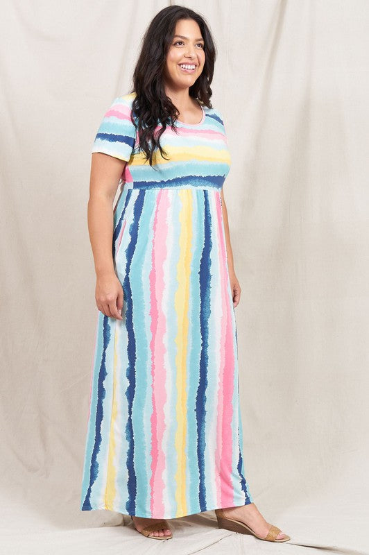 A woman stands against a beige backdrop, wearing the vibrant Water Color Short Sleeve Maxi Dress and holding a woven handbag. She has one hand in her dress pocket and is smiling.