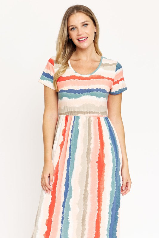 A woman stands against a beige backdrop, wearing the vibrant Water Color Short Sleeve Maxi Dress and holding a woven handbag. She has one hand in her dress pocket and is smiling.