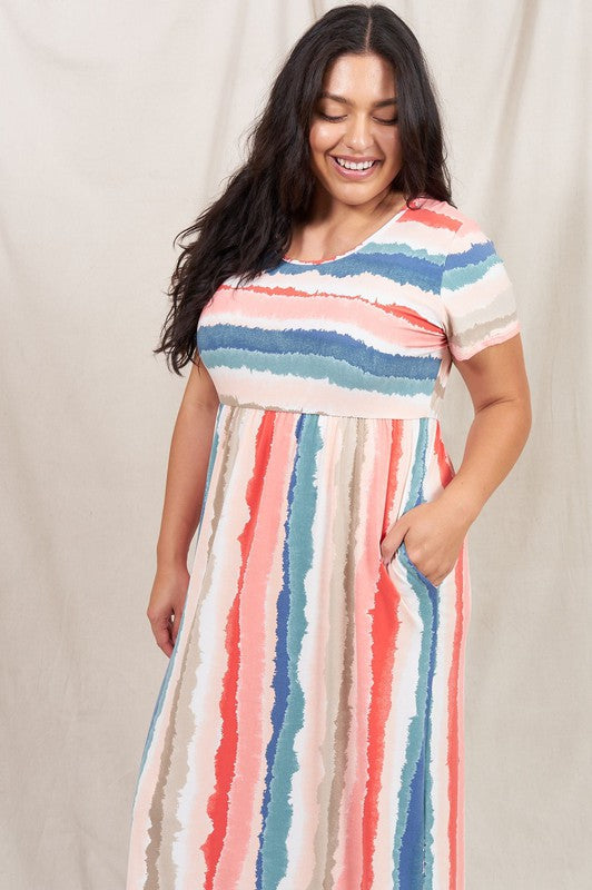 A woman stands against a beige backdrop, wearing the vibrant Water Color Short Sleeve Maxi Dress and holding a woven handbag. She has one hand in her dress pocket and is smiling.