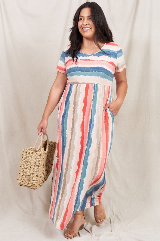 A woman stands against a beige backdrop, wearing the vibrant Water Color Short Sleeve Maxi Dress and holding a woven handbag. She has one hand in her dress pocket and is smiling.