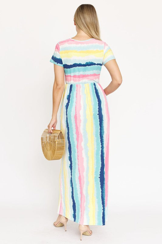 A woman stands against a beige backdrop, wearing the vibrant Water Color Short Sleeve Maxi Dress and holding a woven handbag. She has one hand in her dress pocket and is smiling.