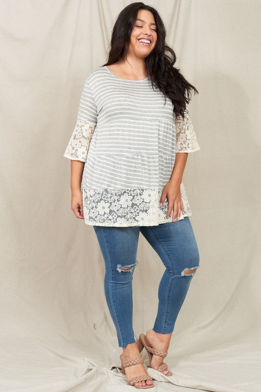 A woman wearing a Plus Lace Trim Tiered Tunic with striped design and lace details on the sleeves and hem stands in front of a plain beige background. She is also wearing ripped jeans.