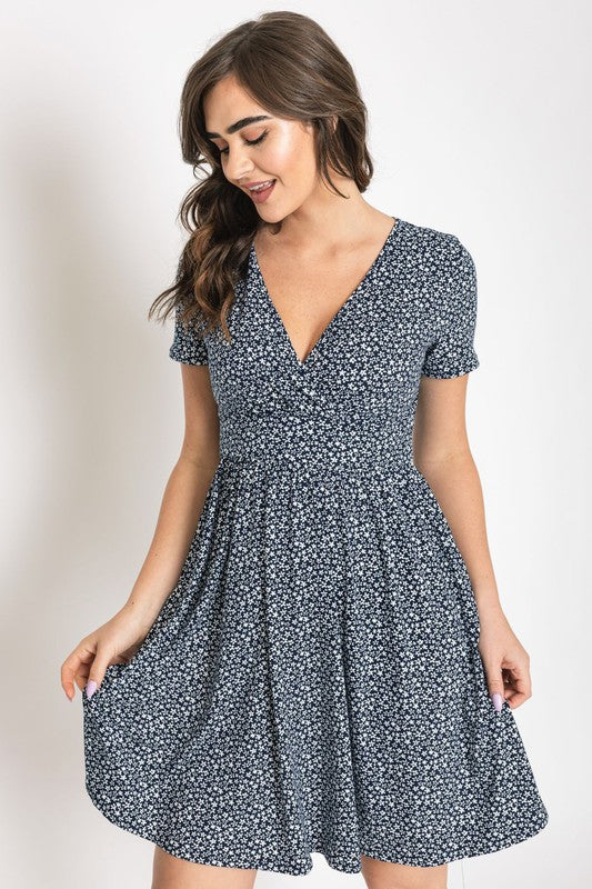 A woman wearing a short-sleeved, V-neck, Ditsy Surplice Fit and Flare Midi Dress with a dark floral background, crafted from luxurious Polyester Spandex and proudly Made in the United States, poses against a white backdrop, smiling as she softly holds the sides of her dress.