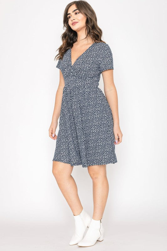 A woman wearing a short-sleeved, V-neck, Ditsy Surplice Fit and Flare Midi Dress with a dark floral background, crafted from luxurious Polyester Spandex and proudly Made in the United States, poses against a white backdrop, smiling as she softly holds the sides of her dress.