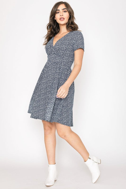 A woman wearing a short-sleeved, V-neck, Ditsy Surplice Fit and Flare Midi Dress with a dark floral background, crafted from luxurious Polyester Spandex and proudly Made in the United States, poses against a white backdrop, smiling as she softly holds the sides of her dress.