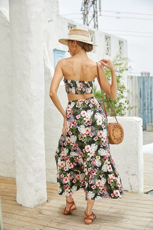 A woman stands on a wooden deck wearing a straw hat and the Women's 2Pcs Floral Long Skirt Set, featuring a chic floral crop top and matching floral long skirt. She complements her stylish ensemble of women's clothing with a brown shoulder bag and brown sandals.