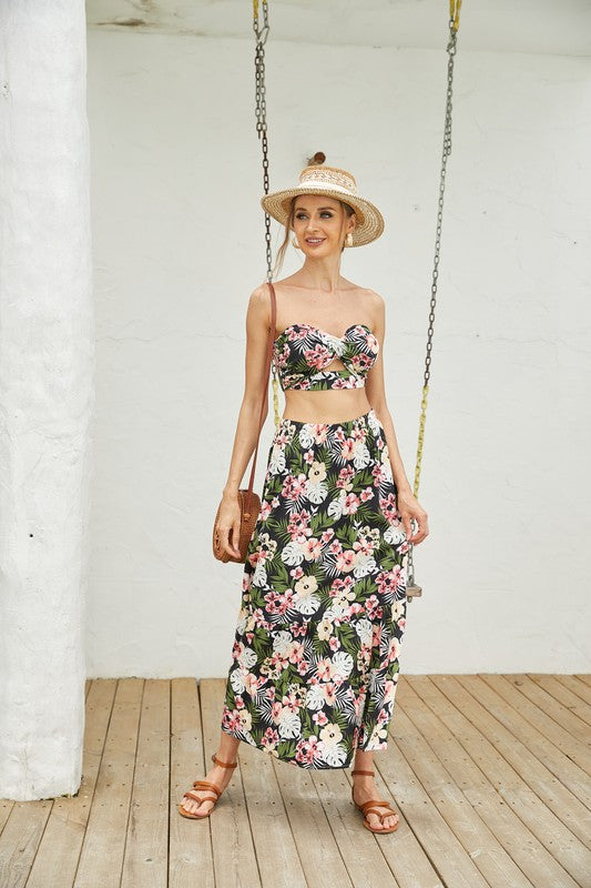 A woman stands on a wooden deck wearing a straw hat and the Women's 2Pcs Floral Long Skirt Set, featuring a chic floral crop top and matching floral long skirt. She complements her stylish ensemble of women's clothing with a brown shoulder bag and brown sandals.