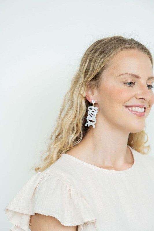 Bride earrings paired with a gold-capped champagne bottle, featuring elegant white script, ideal for a bridal shower and beautifully placed on a white fabric background.