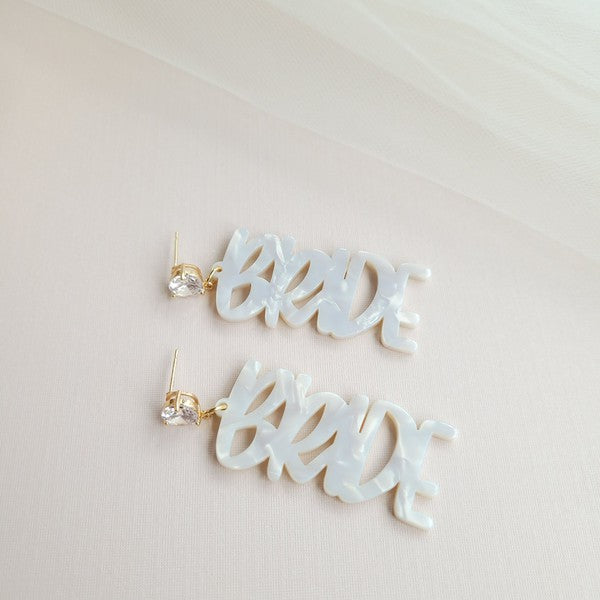 Bride earrings paired with a gold-capped champagne bottle, featuring elegant white script, ideal for a bridal shower and beautifully placed on a white fabric background.