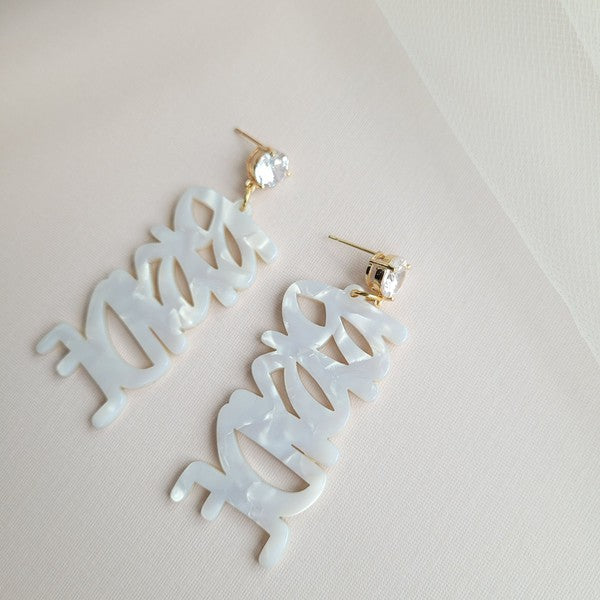 Bride earrings paired with a gold-capped champagne bottle, featuring elegant white script, ideal for a bridal shower and beautifully placed on a white fabric background.