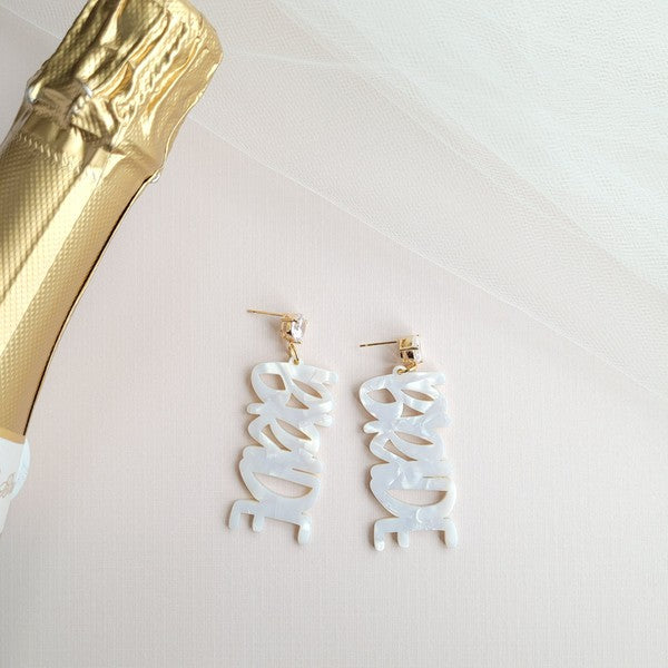 Bride earrings paired with a gold-capped champagne bottle, featuring elegant white script, ideal for a bridal shower and beautifully placed on a white fabric background.