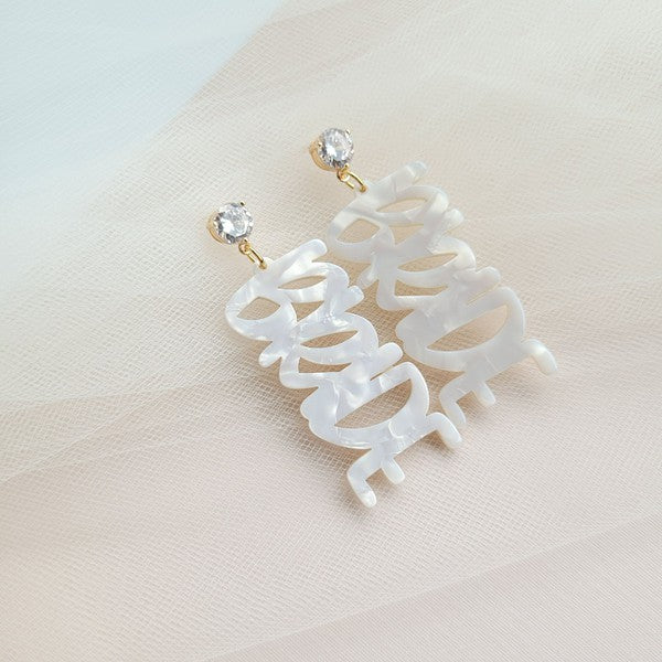 Bride earrings paired with a gold-capped champagne bottle, featuring elegant white script, ideal for a bridal shower and beautifully placed on a white fabric background.