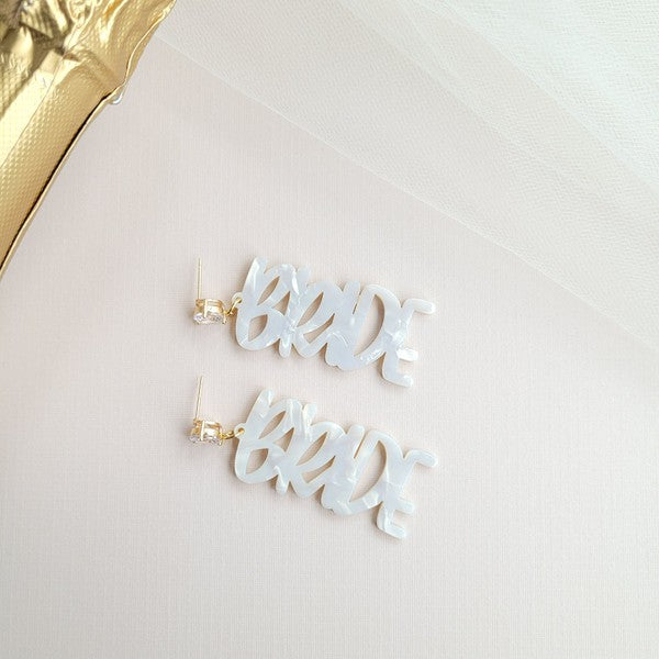 Bride earrings paired with a gold-capped champagne bottle, featuring elegant white script, ideal for a bridal shower and beautifully placed on a white fabric background.