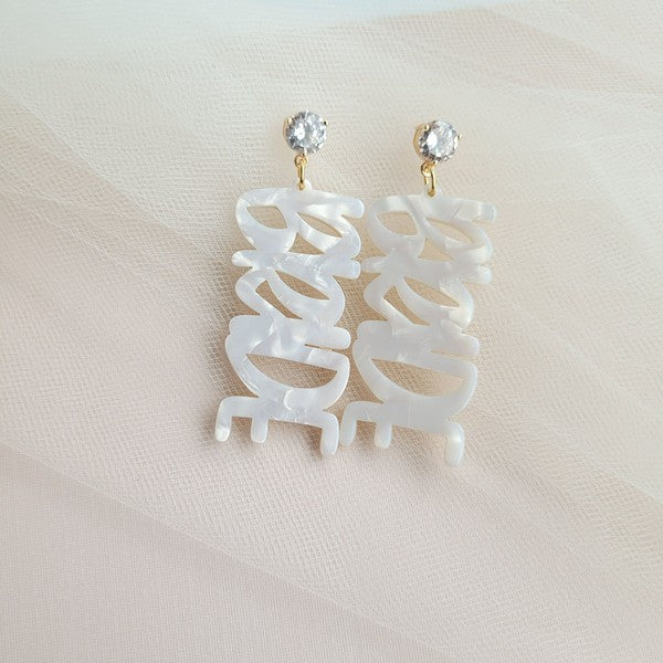 Bride earrings paired with a gold-capped champagne bottle, featuring elegant white script, ideal for a bridal shower and beautifully placed on a white fabric background.